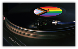 A close-up of a vinyl record spinning on a turntable. The record’s label features the Progress Pride Flag, showing rainbow stripes, chevron, and trans colors. The turntable brand “Technics” is visible in the lower left corner.