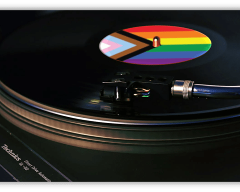 A close-up of a vinyl record spinning on a turntable. The record’s label features the Progress Pride Flag, showing rainbow stripes, chevron, and trans colors. The turntable brand “Technics” is visible in the lower left corner.