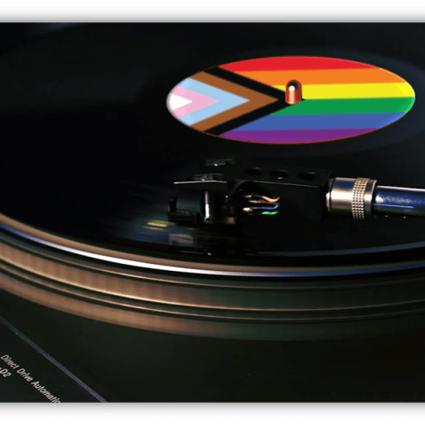 A close-up of a vinyl record spinning on a turntable. The record’s label features the Progress Pride Flag, showing rainbow stripes, chevron, and trans colors. The turntable brand “Technics” is visible in the lower left corner.