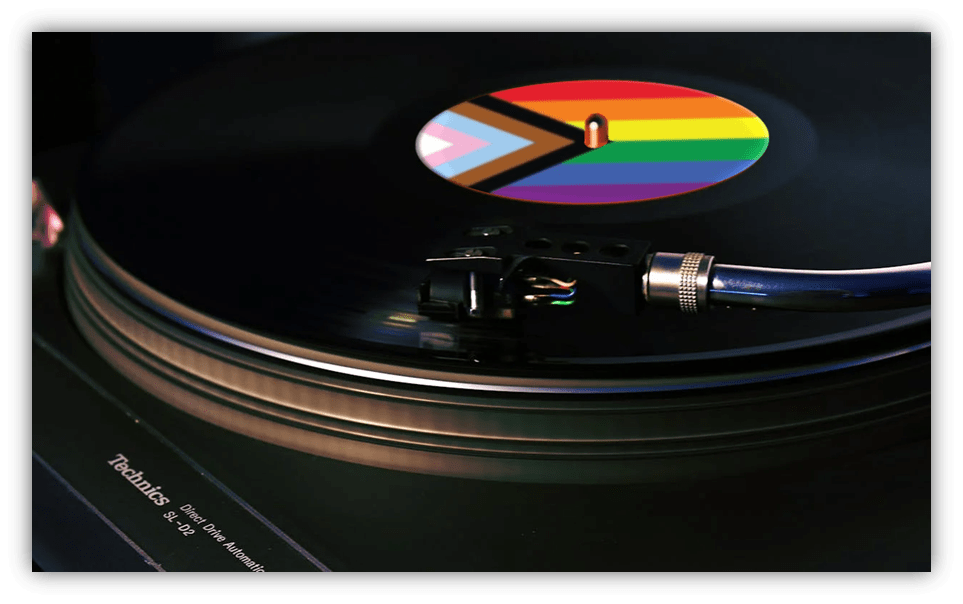 A close-up of a vinyl record spinning on a turntable. The record’s label features the Progress Pride Flag, showing rainbow stripes, chevron, and trans colors. The turntable brand “Technics” is visible in the lower left corner.