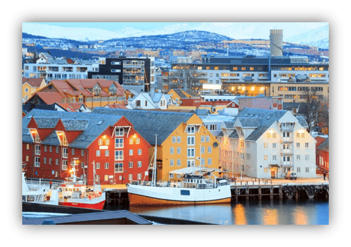 Colorful waterfront buildings in Tromsø, Norway, with fishing boats docked at the harbor, surrounded by snow-capped mountains and a clear blue sky.