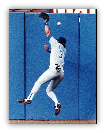 "Baseball player jumping to catch a fly ball against a blue outfield wall."
