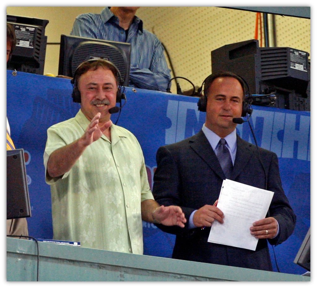 Sports commentators smiling and waving from the broadcast booth.