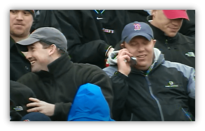 Two men laughing in a crowd, one on a phone, wearing jackets and caps.