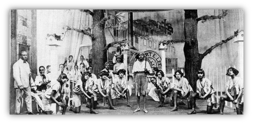 Historical photo of a diverse group of performers in period costumes, staged in front of a backdrop with trees and buildings.