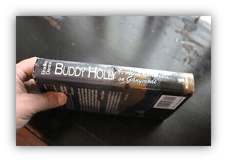 "Person holding a book titled 'Buddy Holly: To Fill the Void on Ganymede' on a dark surface."
