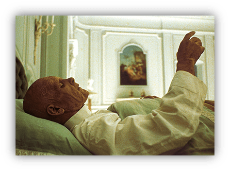 Elderly man lying in bed, gesturing thoughtfully in a softly lit room.