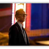 Man in a suit with glasses standing in front of a brightly lit retro sign at night.