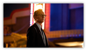 Man in a suit with glasses standing in front of a brightly lit retro sign at night.
