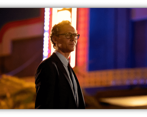 Man in a suit with glasses standing in front of a brightly lit retro sign at night.