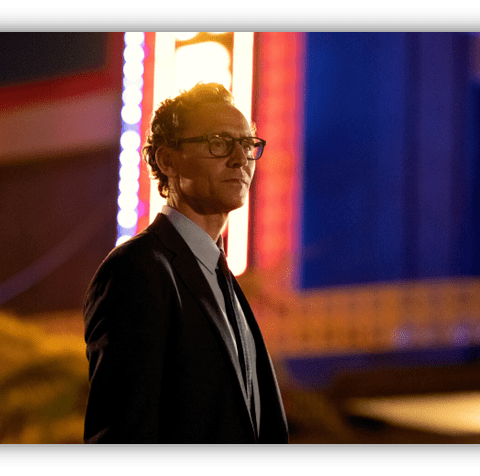 Man in a suit with glasses standing in front of a brightly lit retro sign at night.