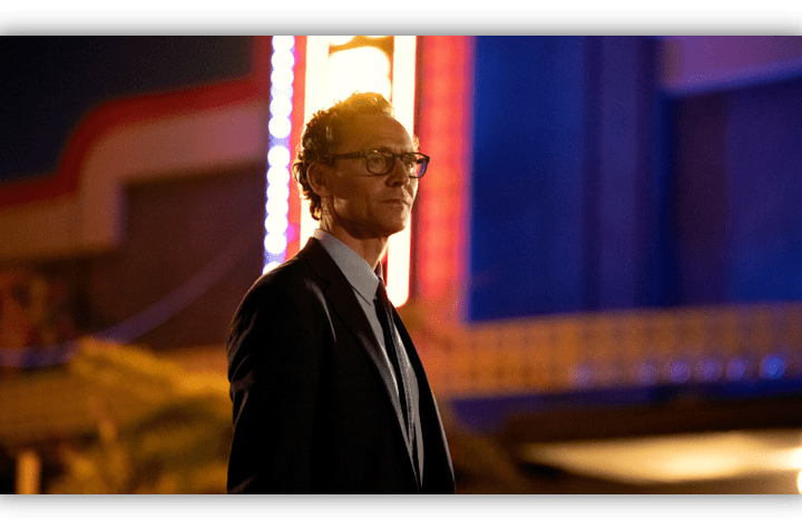 Man in a suit with glasses standing in front of a brightly lit retro sign at night.