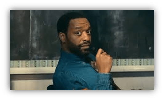 "Man in denim shirt looking back, seated in a classroom setting."