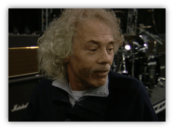 Musician with curly hair speaking in front of a drum set and amplifier.