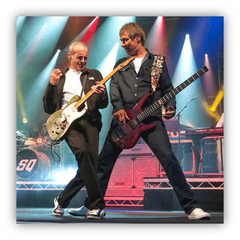 "Two musicians performing on stage with electric guitar and bass, vibrant lights in the background."