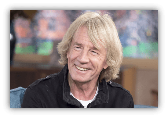 Smiling man with long blonde hair in a black jacket, seated in a studio setting.