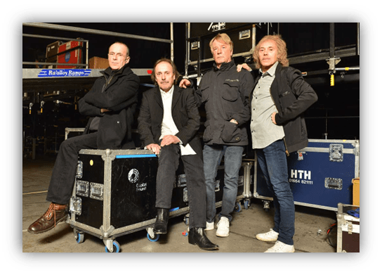 Four musicians posing together in a backstage setting, standing by equipment cases.