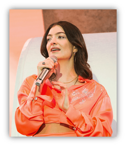 Lorde performing live in an orange outfit, microphone in hand, engaging with the audience.