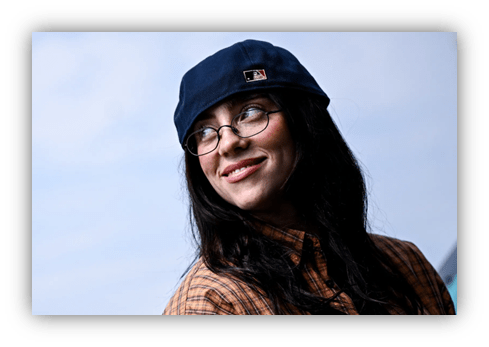 "Smiling woman in a blue cap and glasses, wearing a plaid shirt against a cloudy sky."
