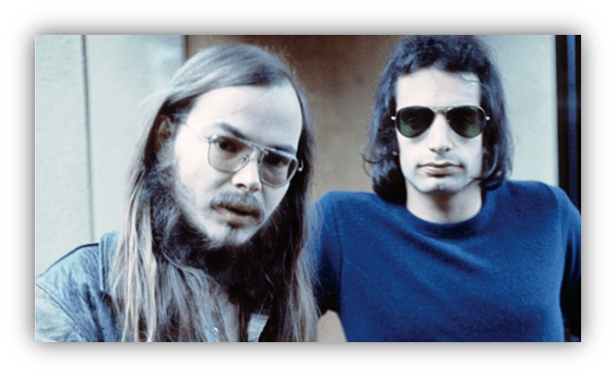 Two men with sunglasses, one with long hair, posing together outdoors.