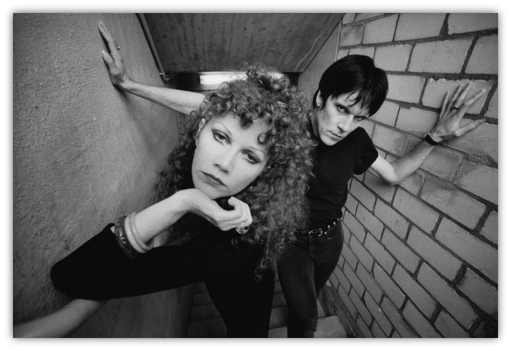 Black and white photo of a couple posing in a stairwell, showcasing edgy fashion and dramatic expressions.