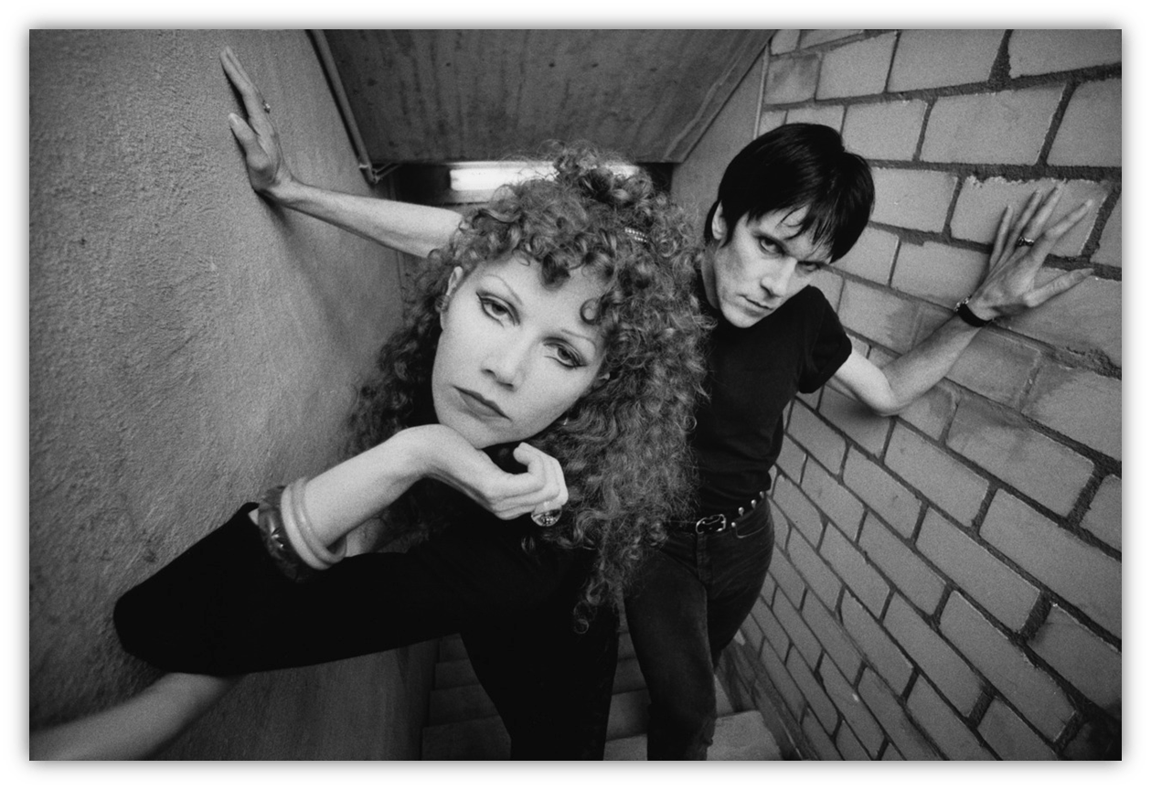 Black and white photo of a couple posing in a stairwell, showcasing edgy fashion and dramatic expressions.