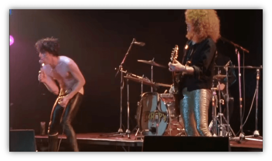 "Live rock performance with a shirtless singer and guitarist in shiny pants."