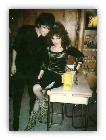 "Two individuals in creative attire pose together at a table, showcasing unique fashion styles."