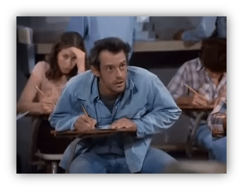 "Man in blue shirt focused on exam in classroom, with students writing in background."