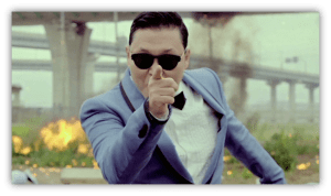 "Man in sunglasses pointing dramatically, dressed in a blue suit with a bow tie, against a backdrop of explosions."