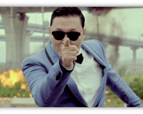 "Man in sunglasses pointing dramatically, dressed in a blue suit with a bow tie, against a backdrop of explosions."