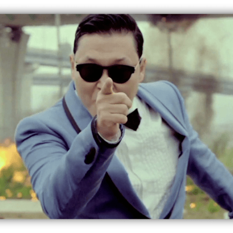 "Man in sunglasses pointing dramatically, dressed in a blue suit with a bow tie, against a backdrop of explosions."