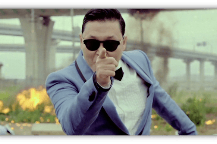 "Man in sunglasses pointing dramatically, dressed in a blue suit with a bow tie, against a backdrop of explosions."