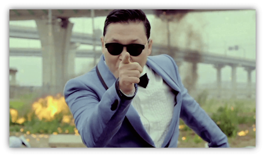 "Man in sunglasses pointing dramatically, dressed in a blue suit with a bow tie, against a backdrop of explosions."