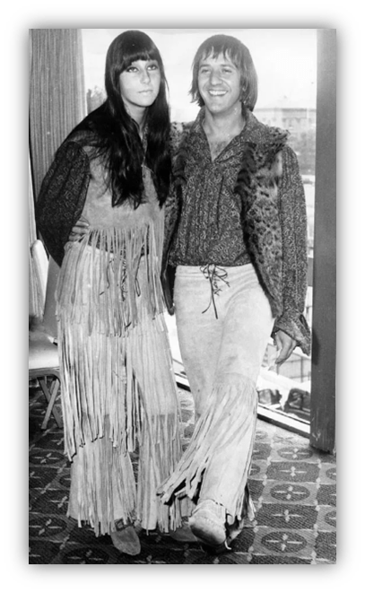 "1970s couple in fringe outfits posing together, showcasing vintage fashion."
