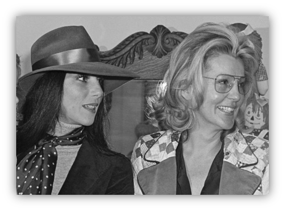 Two women in vintage attire, one in a hat and polka dot scarf, smiling together.
