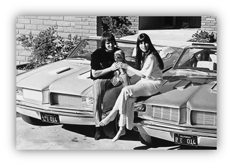 "Two women sitting on classic cars, holding a doll, in a vintage black and white photo."