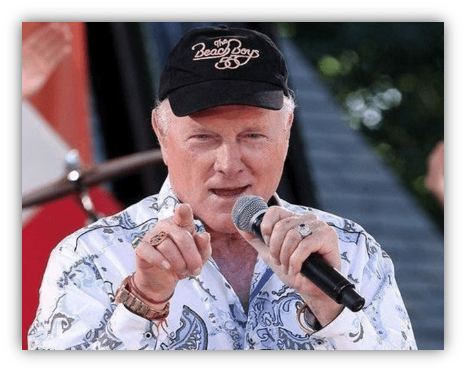 Singer performing on stage, wearing a Beach Boys cap and a patterned shirt, microphone in hand.