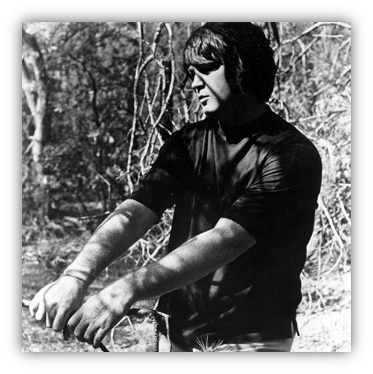 Man standing in a wooded area, holding a stick, in black and white.
