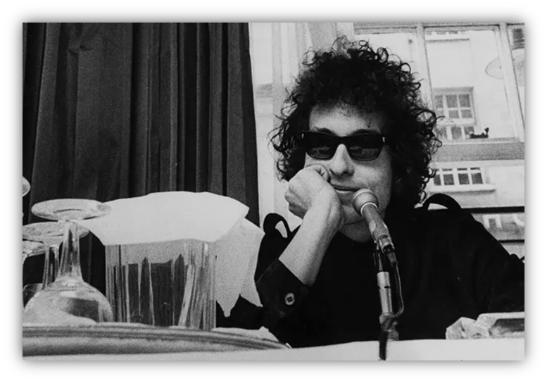"Black and white image of a man with curly hair and sunglasses, resting his chin on his hand at a table with a microphone."
