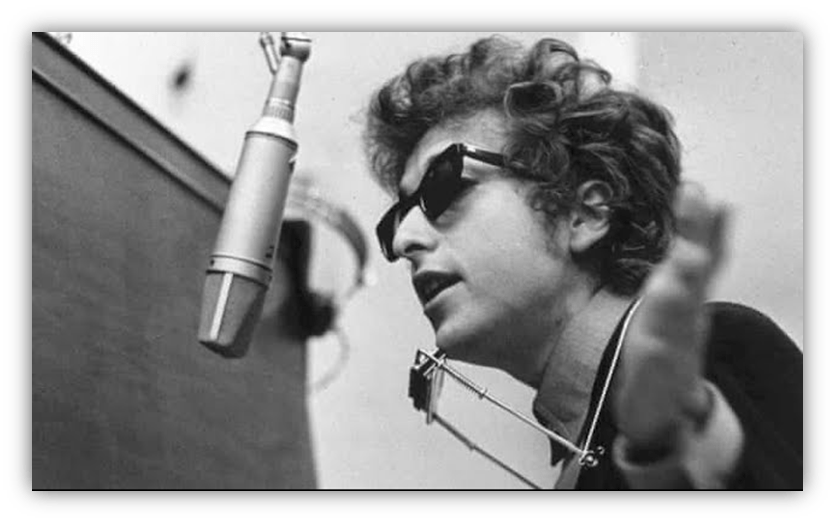"Black and white photo of a singer wearing sunglasses, performing with a harmonica and microphone."
