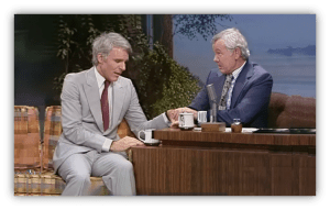 Steve Martin and Johnny Carson share a moment on The Tonight Show, seated at a talk show desk.