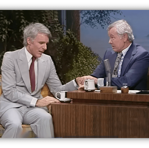Steve Martin and Johnny Carson share a moment on The Tonight Show, seated at a talk show desk.