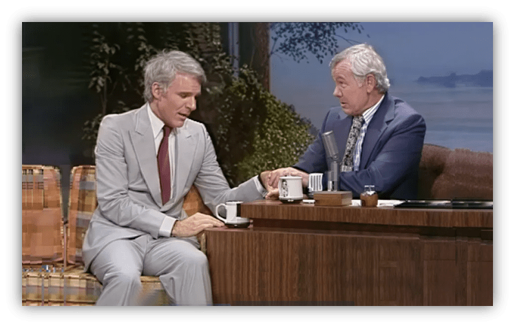 Steve Martin and Johnny Carson share a moment on The Tonight Show, seated at a talk show desk.