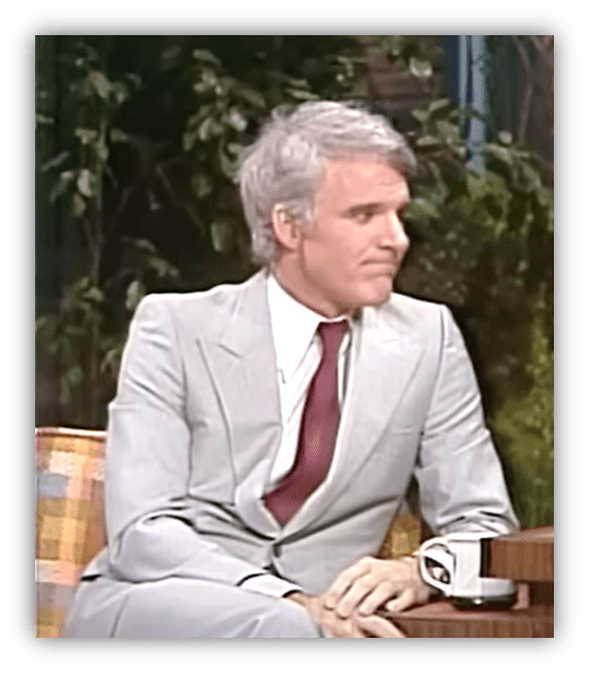 Steve Martin in a light gray suit during a talk show interview.