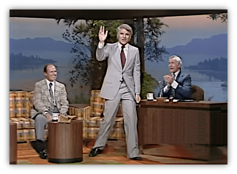Steve Martin waves on stage during a classic talk show, with two hosts applauding.