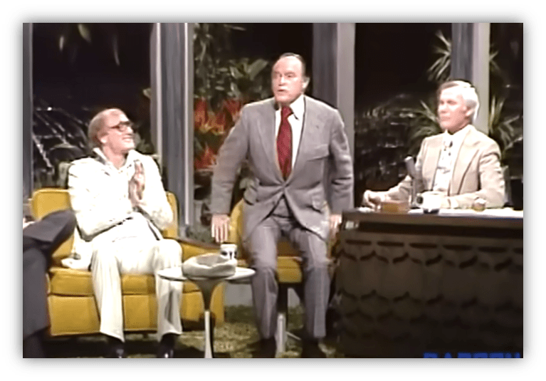 "Classic talk show scene featuring three male hosts, with one standing and two seated, amid a tropical backdrop."