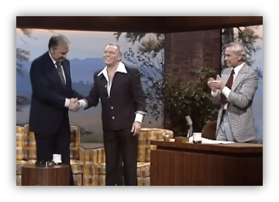 "Two men shaking hands on a talk show set, with a host applauding in the background."