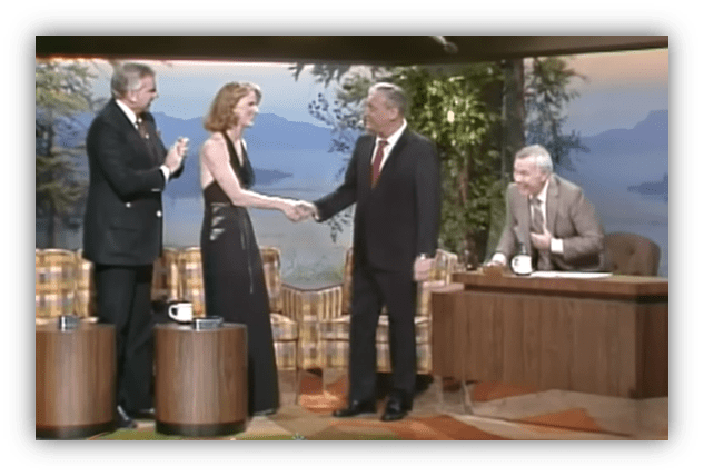 "Talk show hosts greet a guest on stage, with a scenic backdrop."