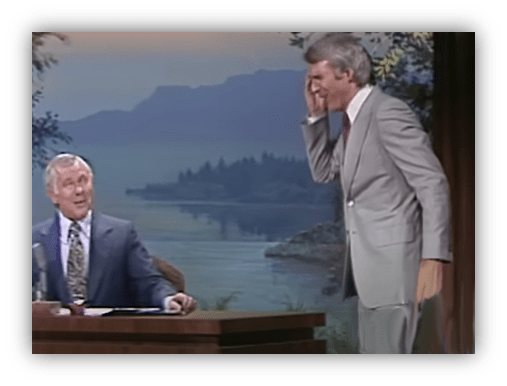 Johnny Carson laughs as a guest gestures playfully on a talk show set.